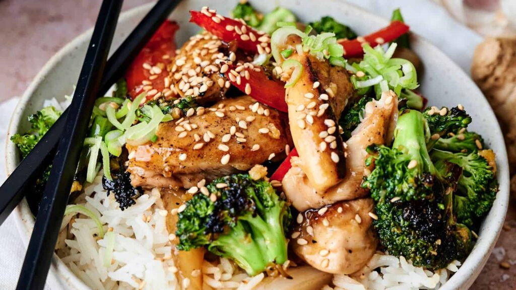 A bowl of rice topped with chicken, broccoli, red bell peppers, and green onions, garnished with sesame seeds. Black chopsticks rest on the bowl's edge.