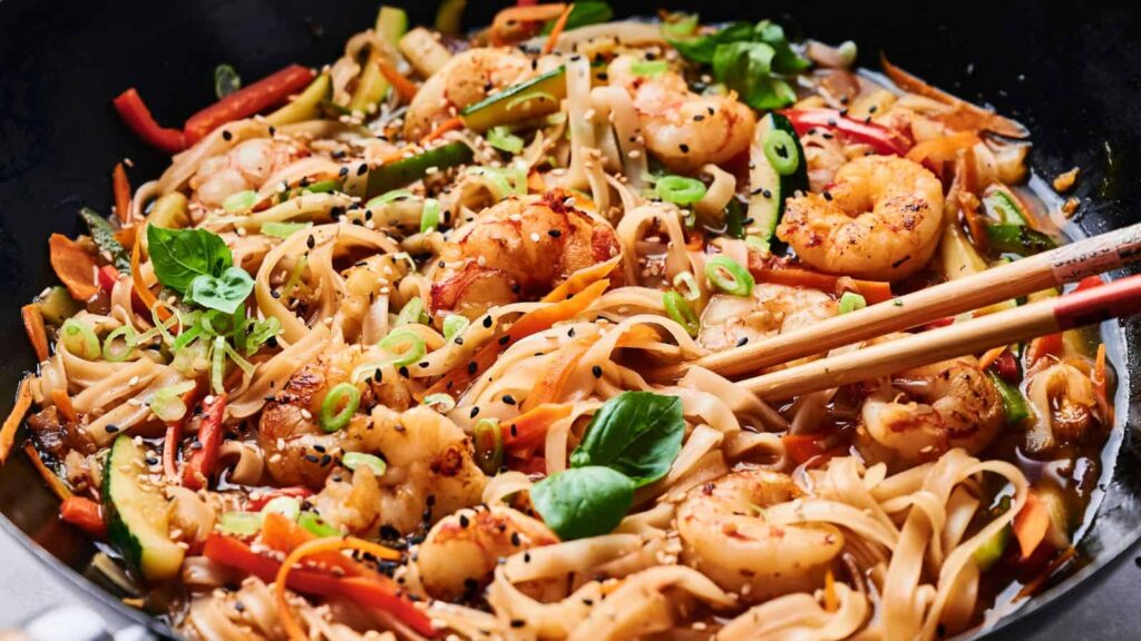 A bowl of shrimp stir-fry noodles with sliced vegetables, garnished with green onions, basil leaves, and sesame seeds, with chopsticks resting on top.