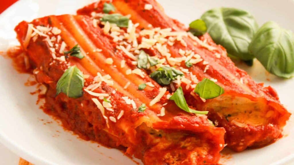 Plate of baked cannelloni filled with cheese, topped with tomato sauce, grated cheese, and fresh basil leaves.