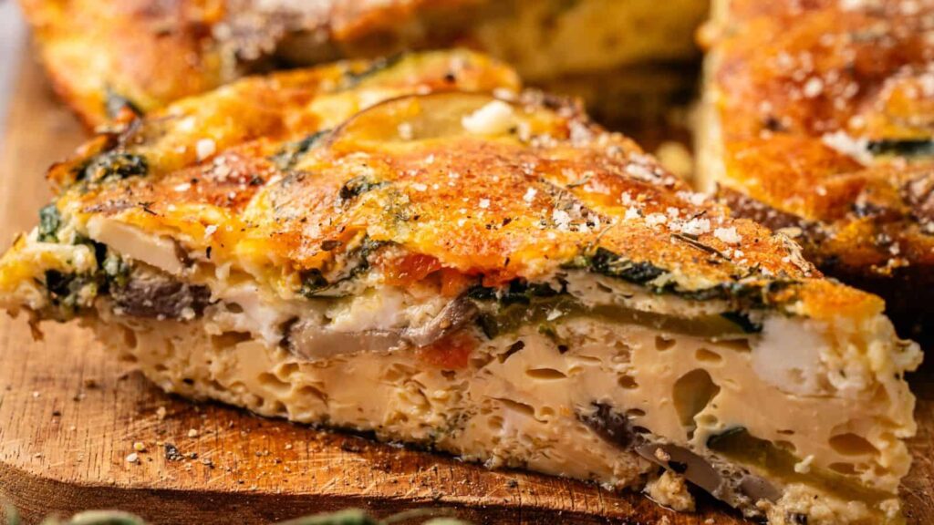 A close-up of a slice of vegetable frittata on a wooden surface, showing layers of egg, vegetables, and seasoning.