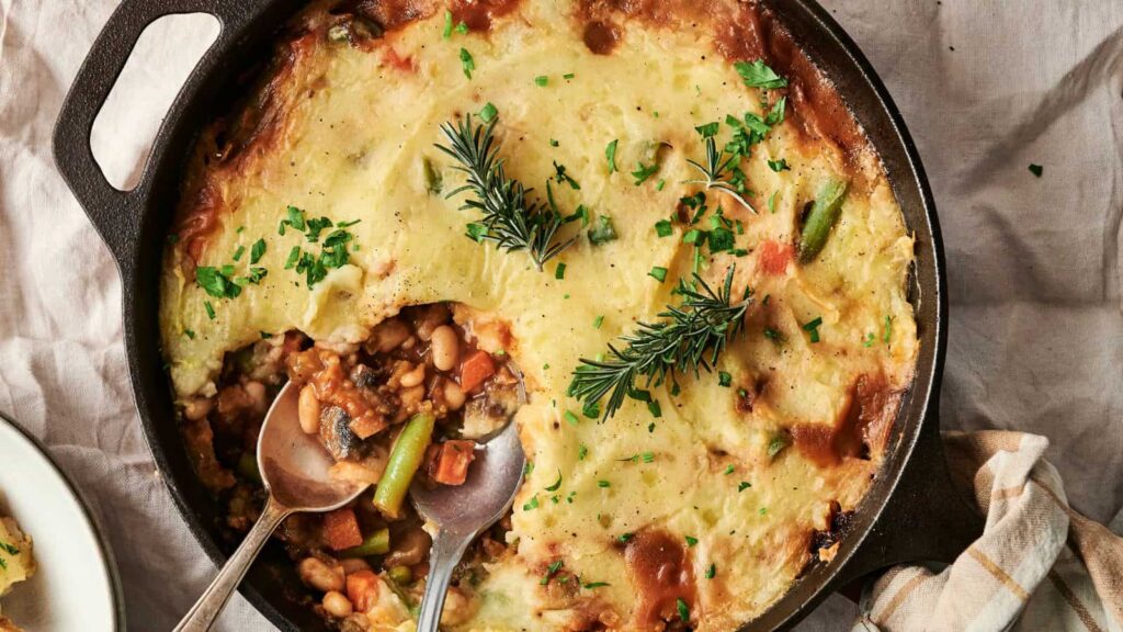 A cast iron skillet with a vegetable and bean shepherd's pie, topped with mashed potatoes and garnished with rosemary and parsley. Two serving spoons are placed in the skillet.