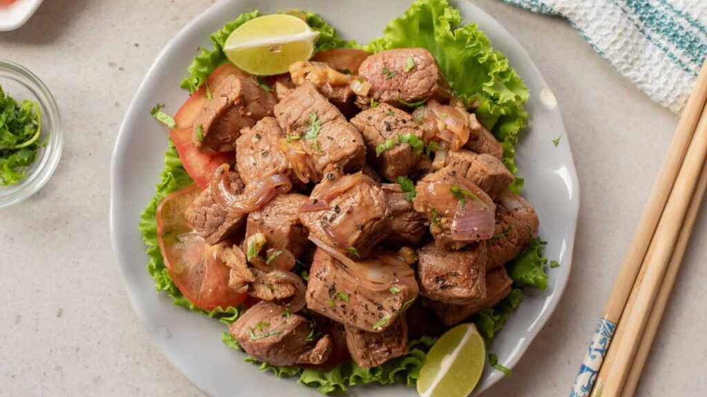 A plate of sautéed beef cubes with onions, garnished with chopped cilantro, lettuce, tomato slices, and lime wedges, served on a white plate with chopsticks beside it.