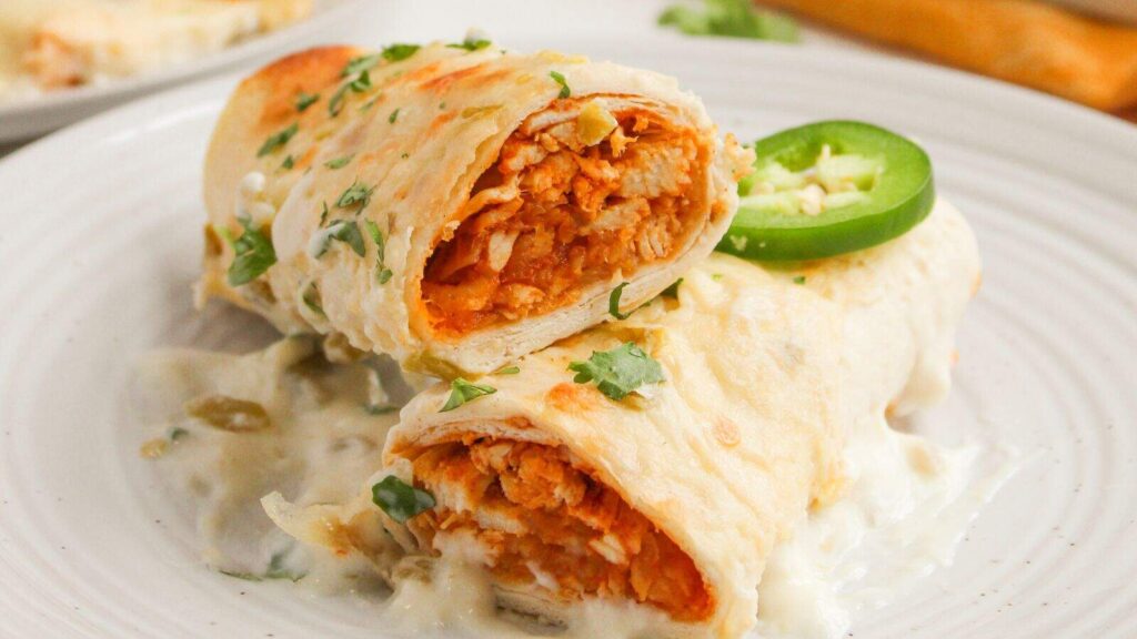 Two tortilla-wrapped enchiladas filled with shredded chicken, topped with creamy white sauce, chopped cilantro, and a jalapeño slice, served on a white plate.