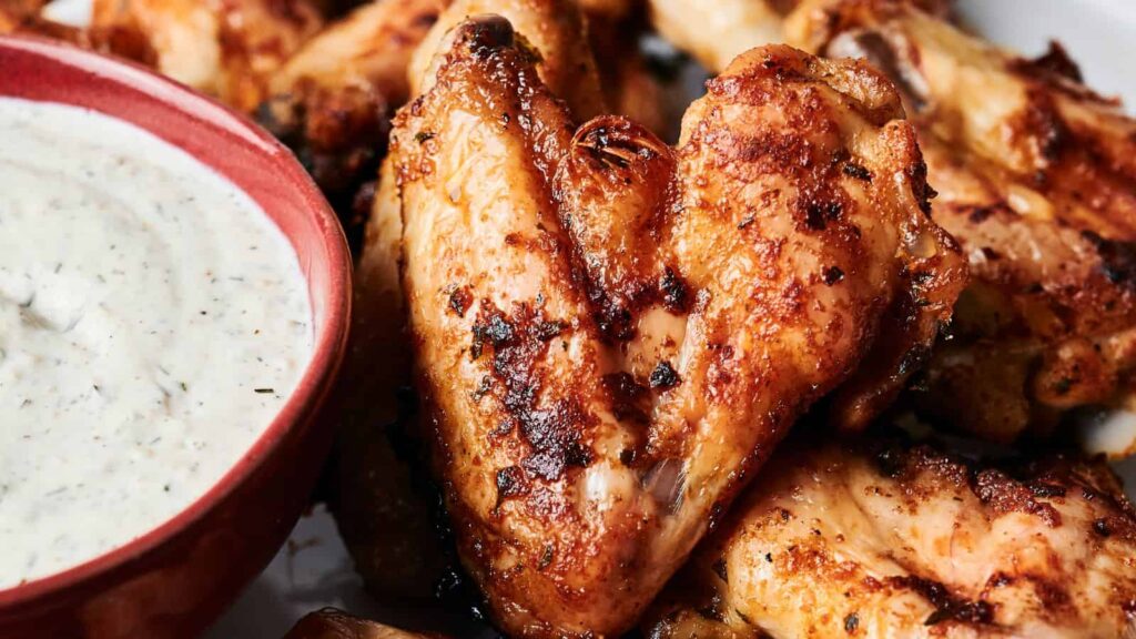 Close-up of grilled chicken wings with a crispy texture next to a red bowl of creamy, white dipping sauce.