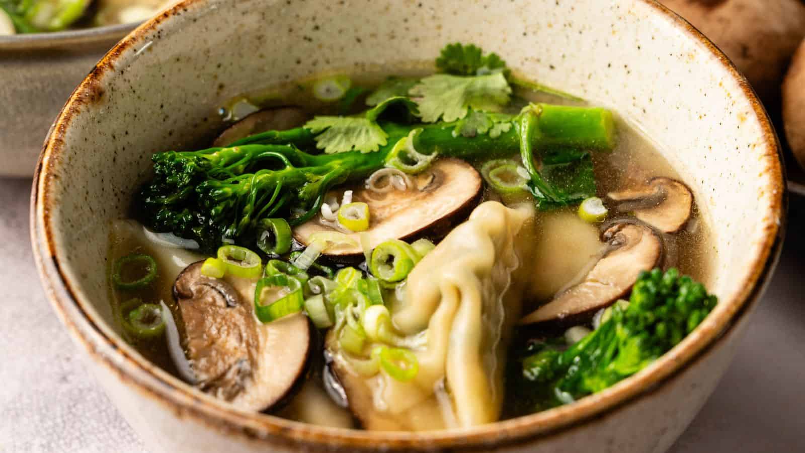 A ceramic bowl of clear soup containing dumplings, sliced mushrooms, broccolini, chopped green onions, and cilantro.