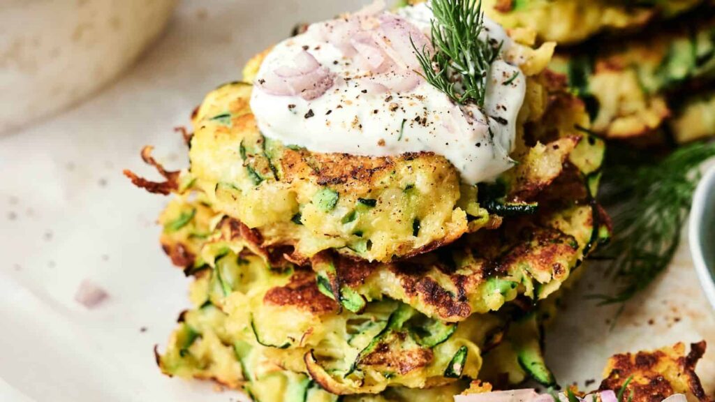 A stack of zucchini fritters topped with dollop of cream, sliced shallots, and dill on a plate.