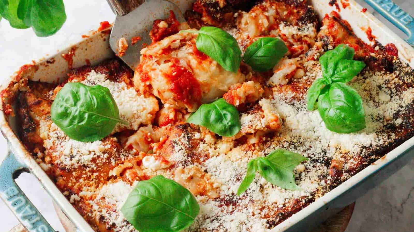 A baked lasagna in a rectangular dish, topped with tomato sauce, basil leaves, and grated cheese. A spatula is lifting a portion from the dish, revealing layers of pasta and filling. The dish is on a light countertop.