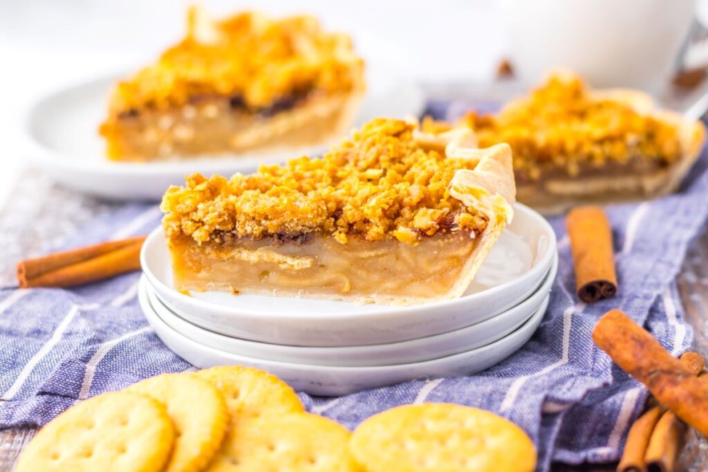 Slice of pie with crumb topping on a stack of plates, surrounded by crackers and cinnamon sticks.
