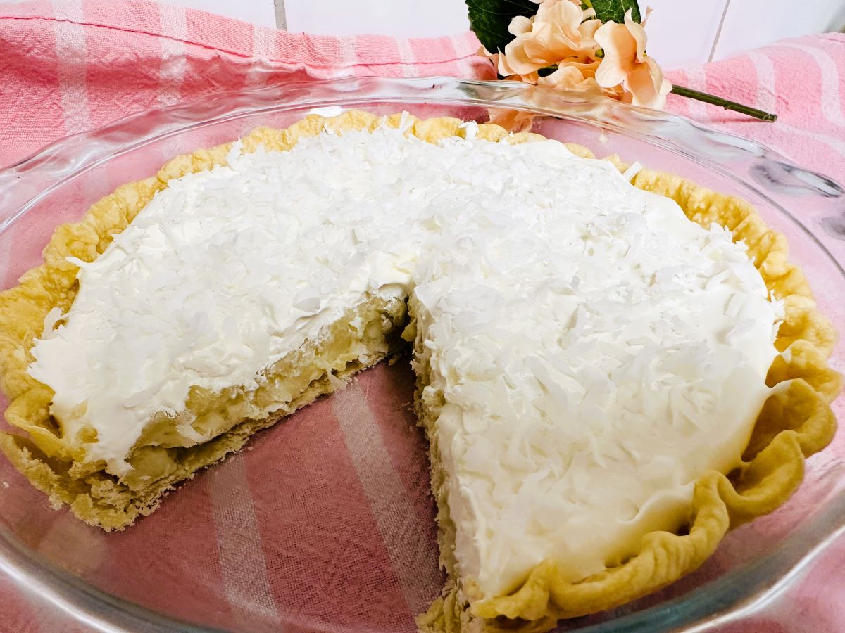 An Old Fashioned Coconut Cream Pie in a glass dish, with a slice missing to reveal its creamy filling and shredded coconut topping. The pie rests on a pink and white striped cloth, peach-colored flowers in the background.