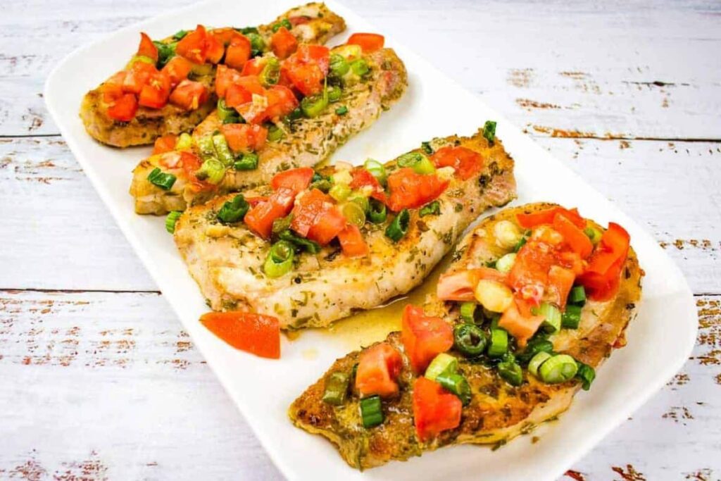 Four grilled chicken breasts garnished with chopped tomatoes and green onions are arranged on a rectangular white plate.