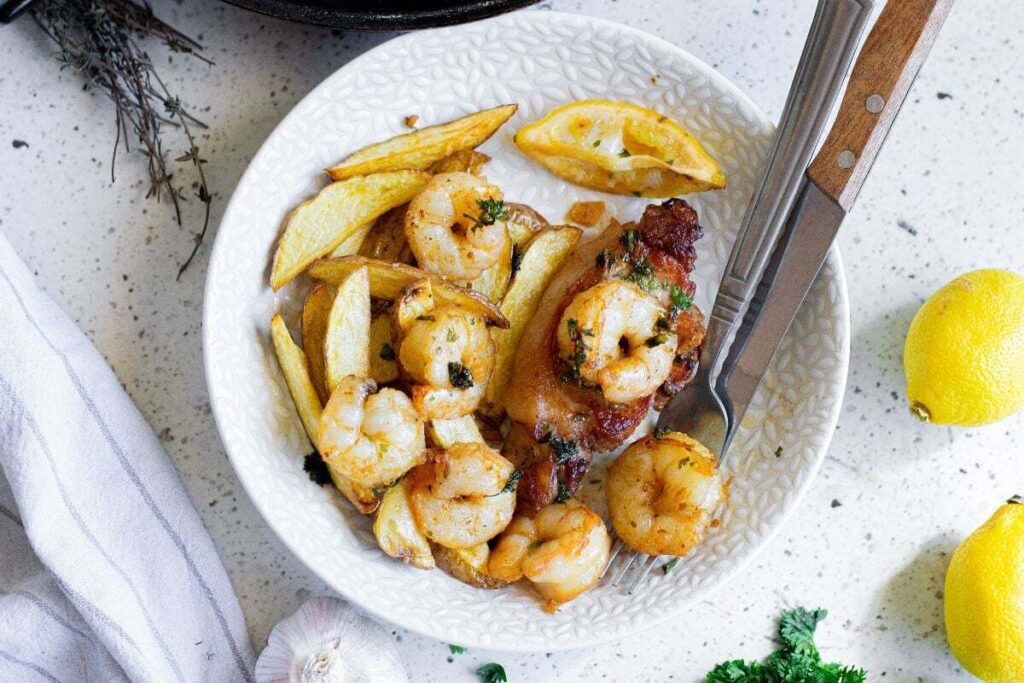 White bowl with buttered herbed shrimp, chicken, potato wedges, and a lemon wedge.