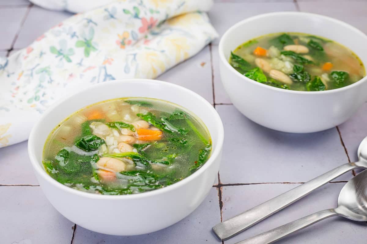Two white bowls of Rice and White Bean Soup on a tiled surface with two metal spoons nearby.