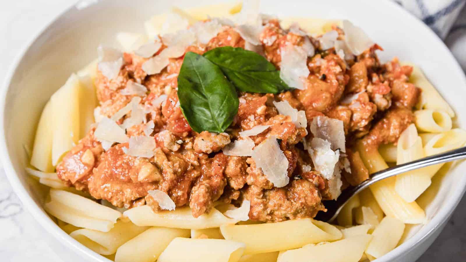 A white bowl filled with penne pasta topped with meat sauce, grated parmesan cheese, and fresh basil leaves, with a metal spoon resting on the side of the bowl.