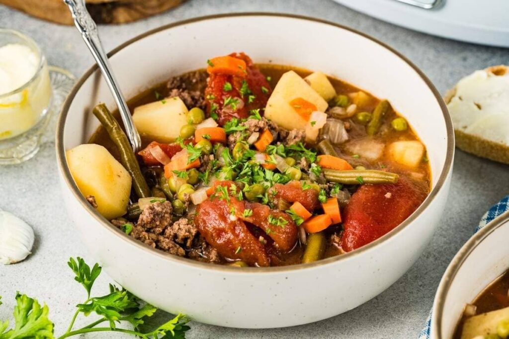 A bowl of slow cooker poor man's stew (hamburger stew), containing chunks of beef, potatoes, green beans, tomatoes, carrots, and peas. It's made effortlessly in a Crockpot and garnished with chopped parsley.