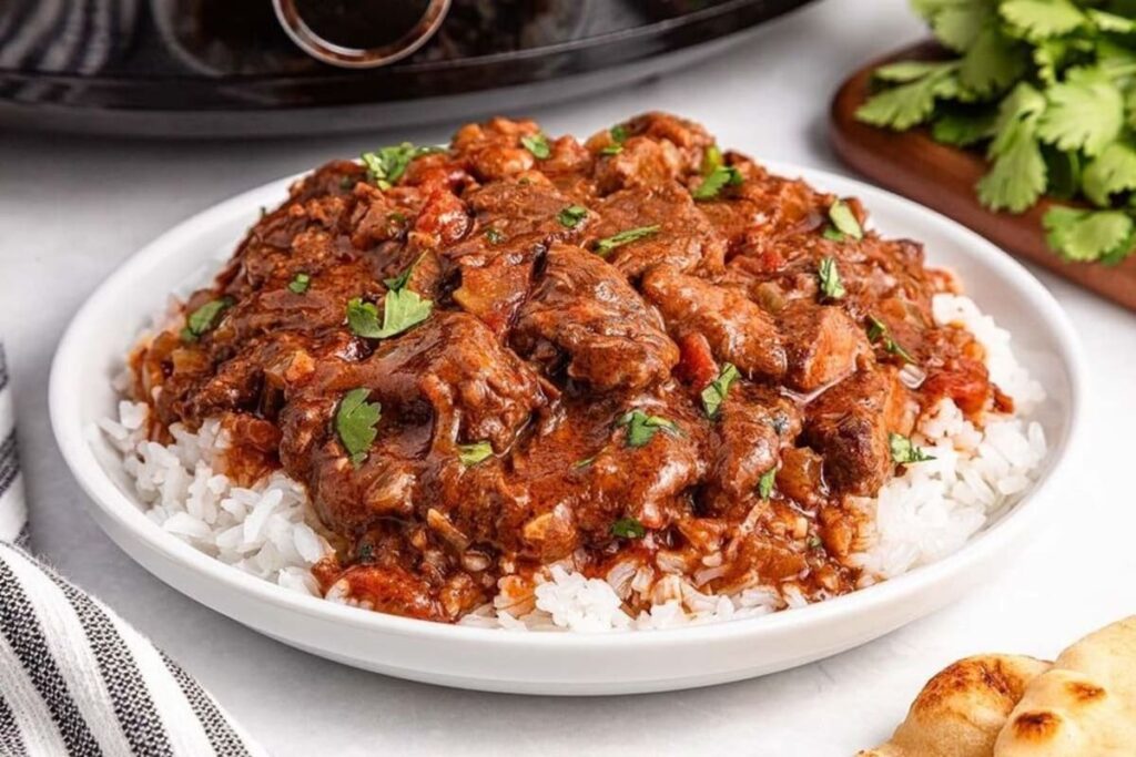 A plate of slow cooker beef curry over rice, garnished with cilantro, sits next to a striped napkin and cilantro bunch.