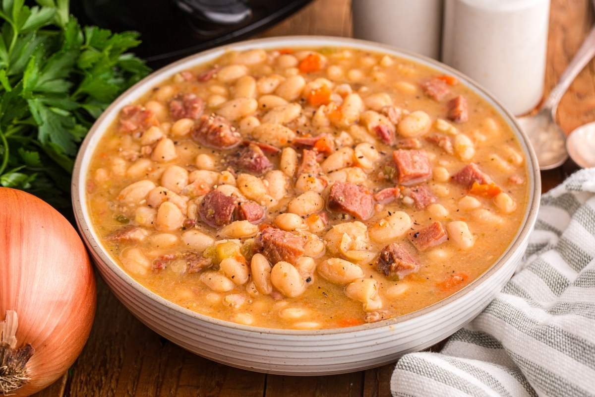 A bowl of Ham and Bean Soup with visible chunks of meat, seasoned with herbs, next to a whole onion and fresh parsley.