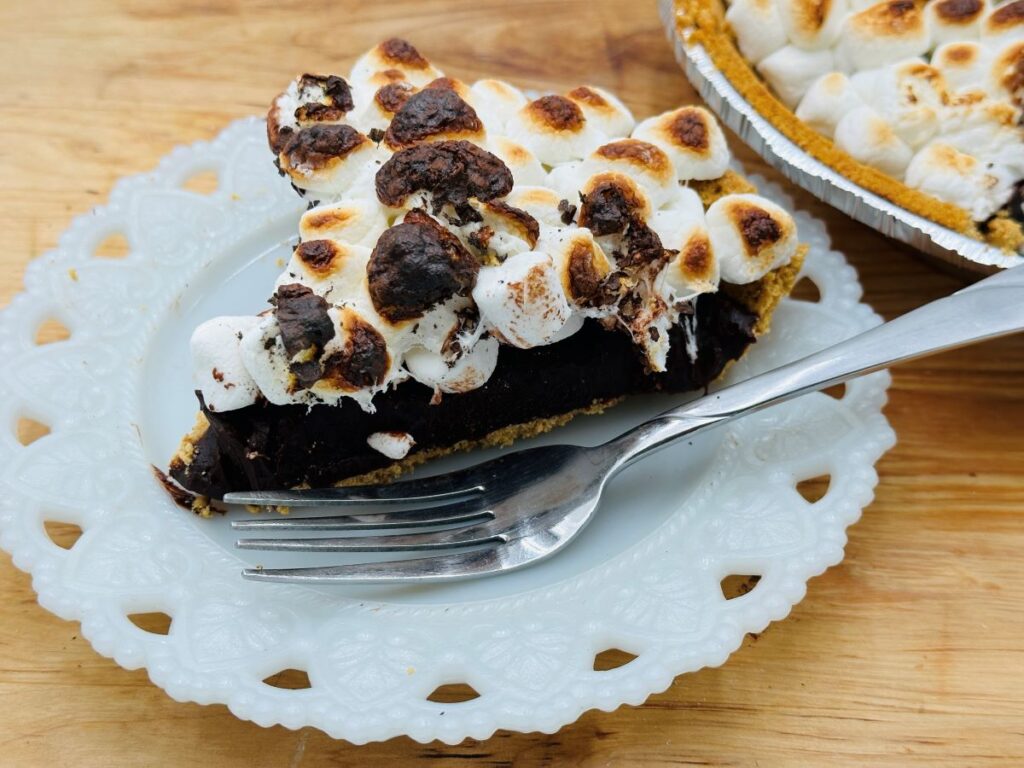 A slice of chocolate pie topped with toasted marshmallows on a white ornate plate, with a fork placed beside it, rests on a wooden surface. Another partially visible pie is in the background.