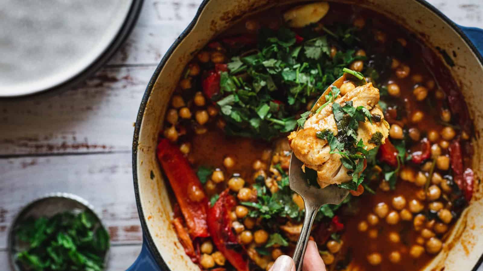 A fork holds a bite of chickpea stew with herbs above a pot filled with stew, chickpeas, and greens.