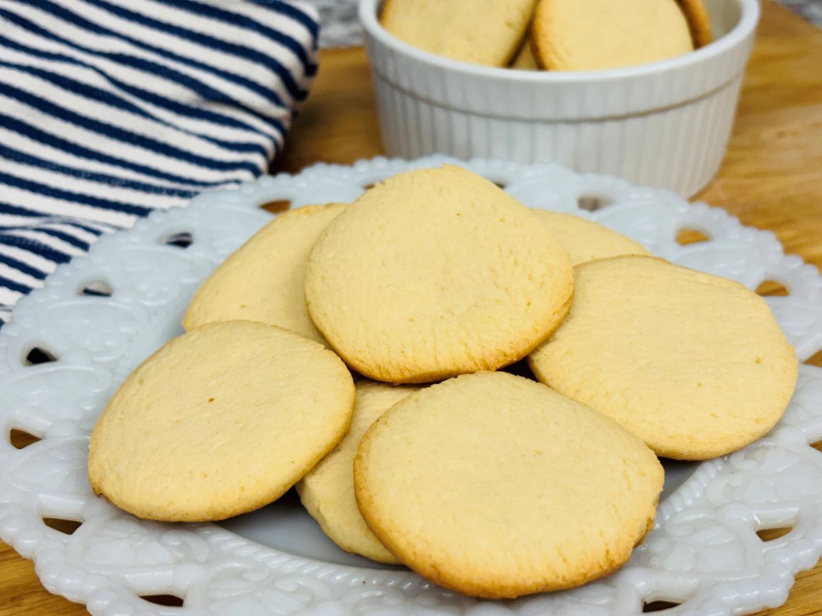 A white plate with round, golden-brown cookies sits on a wooden surface next to a bowl of cookies and a blue-and-white striped cloth.