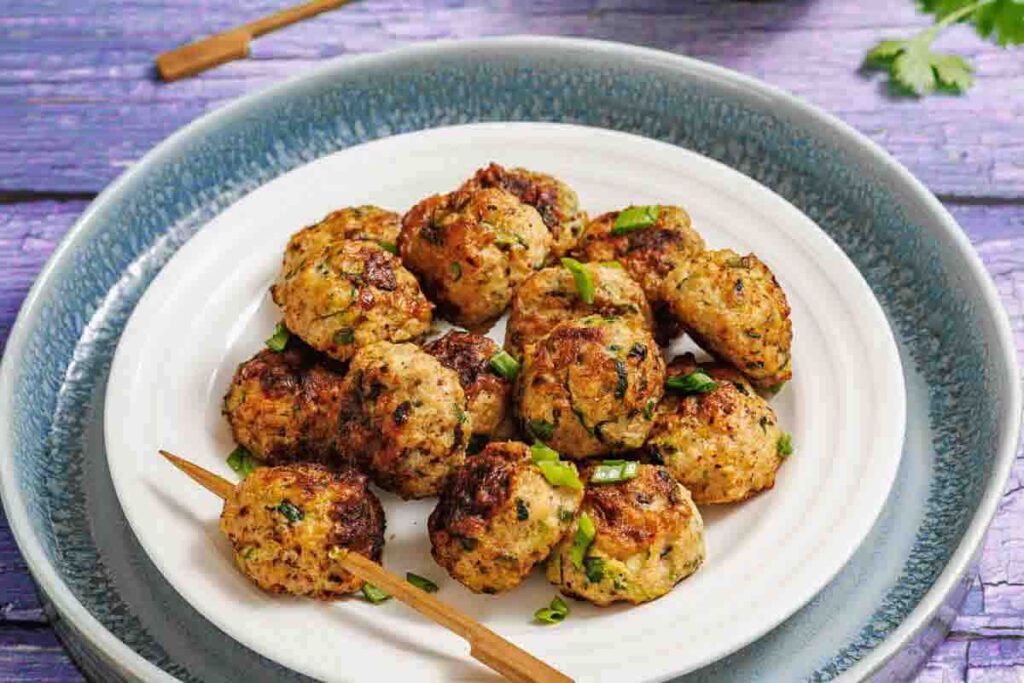A white plate holds a serving of browned, herb-flecked meatballs with two wooden skewers, set on a blue-rimmed plate atop a purple wooden surface.