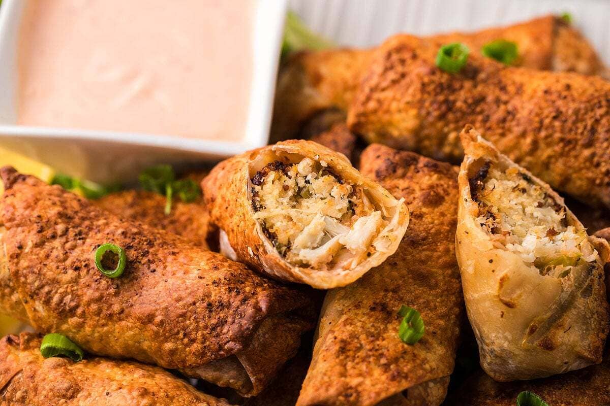 Crispy egg rolls on a plate with chopped green onions, one cut open to show the filling, with a small bowl of dipping sauce nearby.