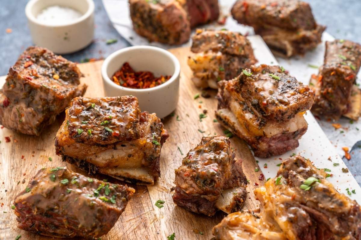 Several pieces of seasoned and cooked ribs on a wooden cutting board, garnished with herbs. Small bowls with spices are placed on the board.