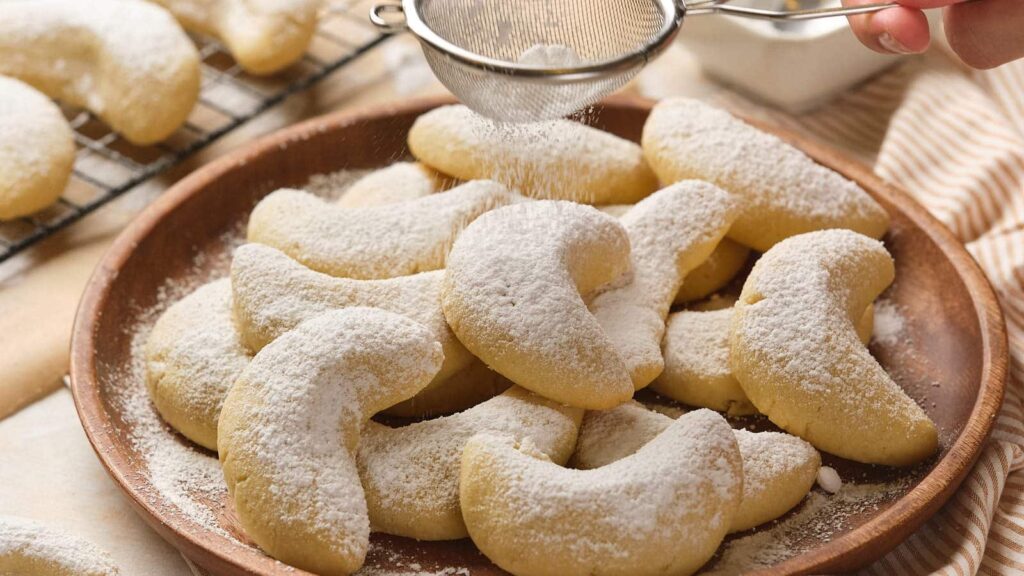 Almond crescent cookies are being dusted with powdered sugar on a wooden plate.