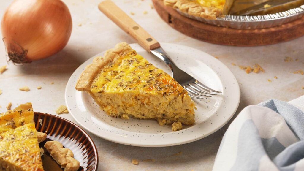 A slice of savory quiche with herbs sits on a white speckled plate.