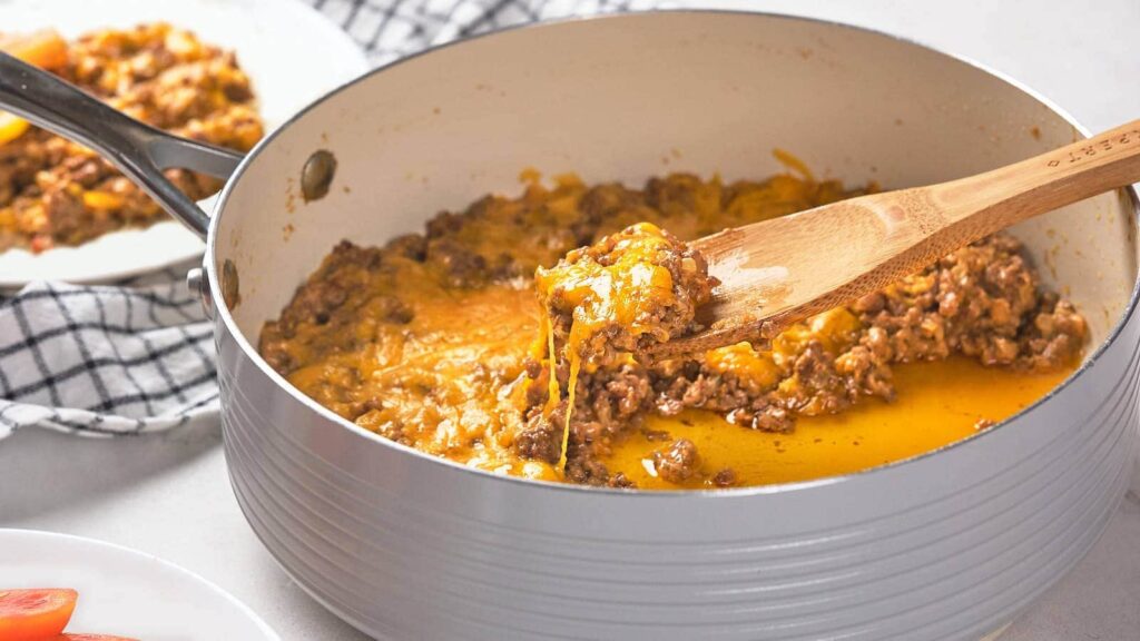A wooden spoon lifting a portion of cheeseburger skillet.