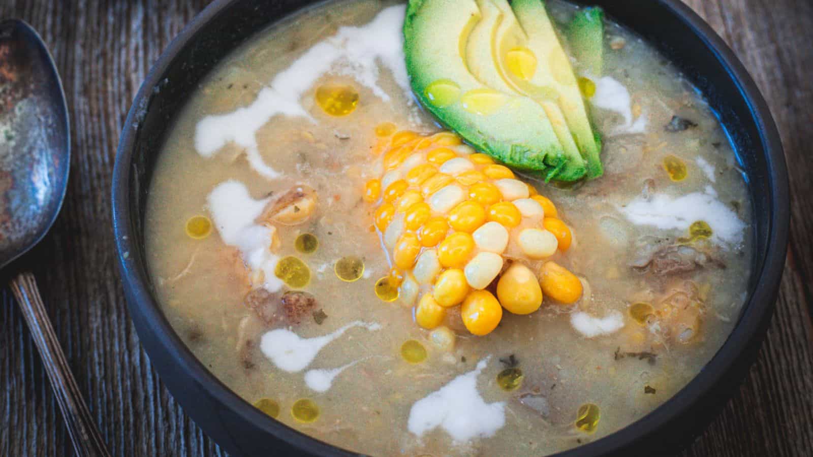 A bowl of ajiaco colombiano soup with chicken, corn on the cob, and avocado.