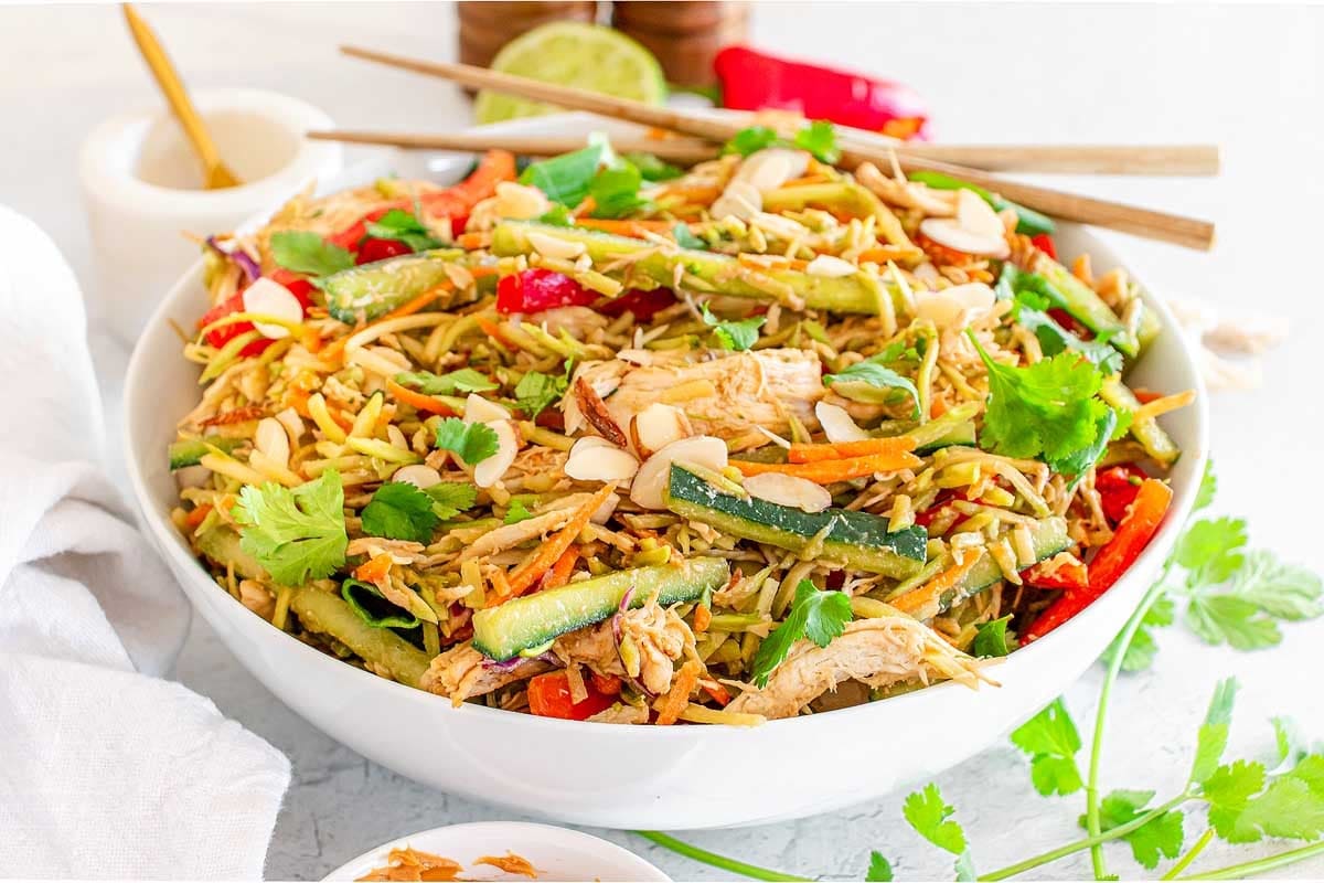 A bowl of Chinese chicken salad with shredded chicken, snap peas, carrots, and peppers, accompanied by a bottle of dressing on the side.