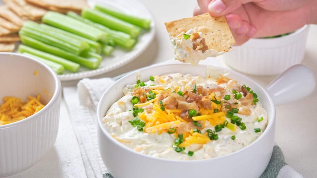 Crack dip in a white bowl with a person's hand dipping a piece of nacho in it.