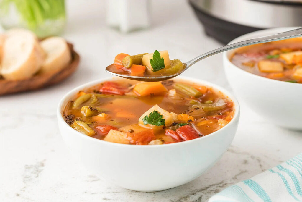 A spoon taking a scoop of vegetable soup out of a white bowl.