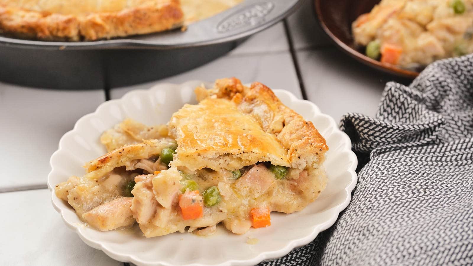 A serving of chicken pie with a golden crust on a white plate, showing pieces of chicken, peas, and carrots, with more pie in the background.
