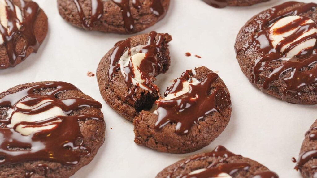 Hot chocolate cookies drizzled with chocolate syrup are resting on a white surface.