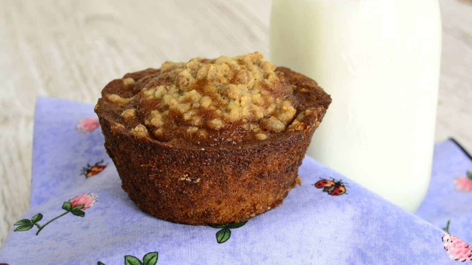 A crumb-topped muffin on a floral napkin exudes the cozy charm of a crisp fall day, perfectly paired with a glass milk bottle.