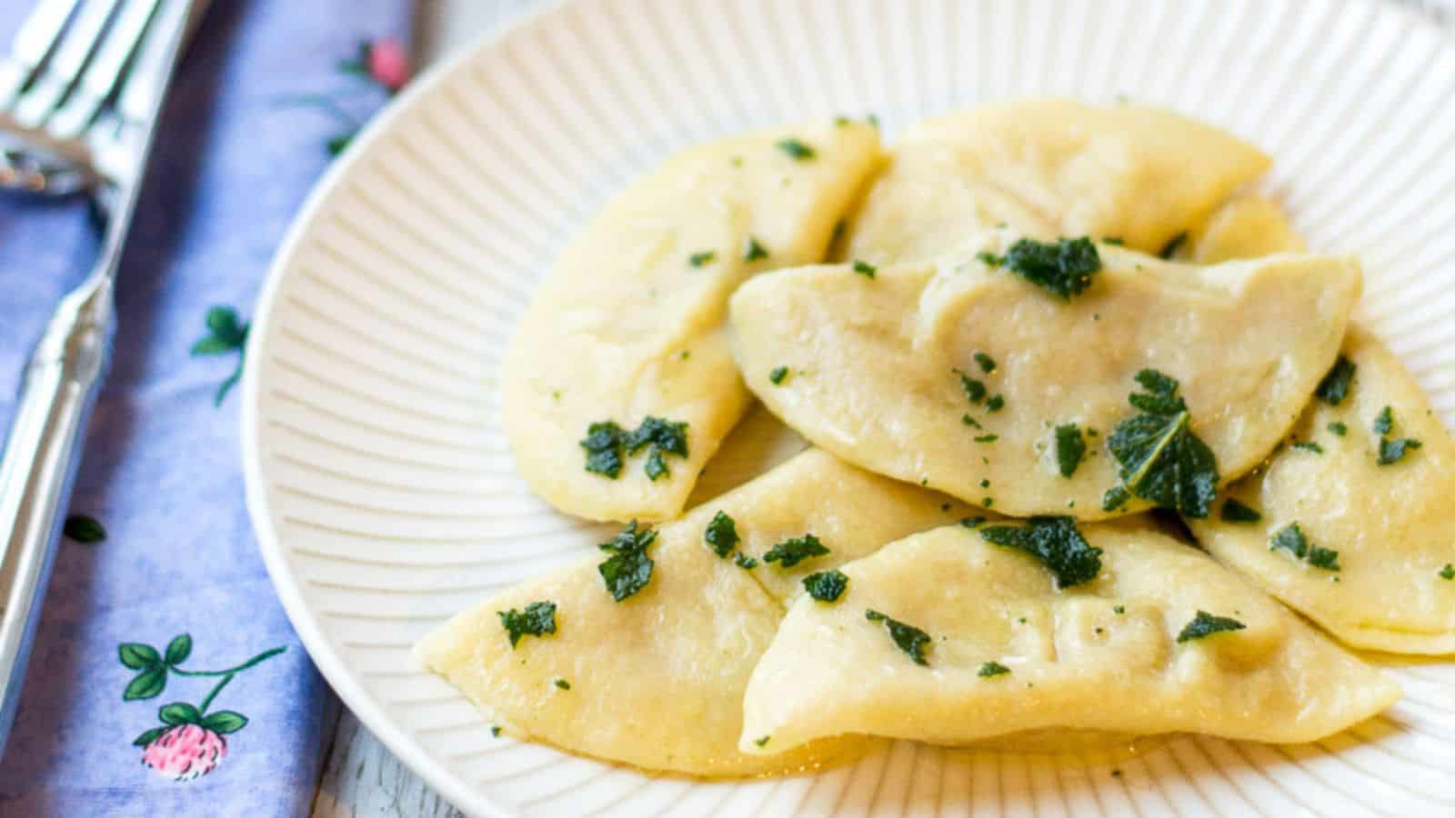 A white plate with six half-moon shaped cooked dumplings garnished with herbs, perfect as a side dish or for kids. A fork and a blue napkin with floral print are placed beside the plate.
