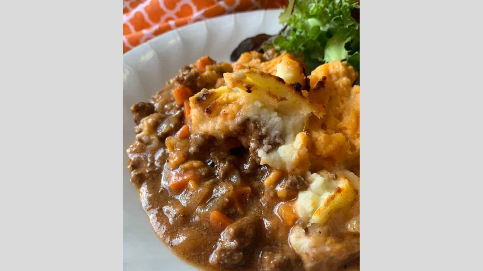 A plate with cottage pie casserole and a side of mixed greens on a white surface, accompanied by an orange-patterned cloth in the background.