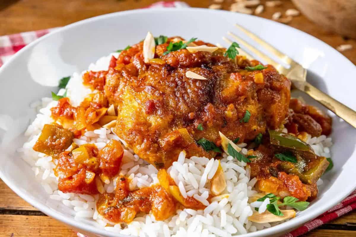 A plate of white rice topped with tender Country Captain chicken in tomato sauce, garnished with chopped parsley and slivered almonds, served with a gold fork on the side.