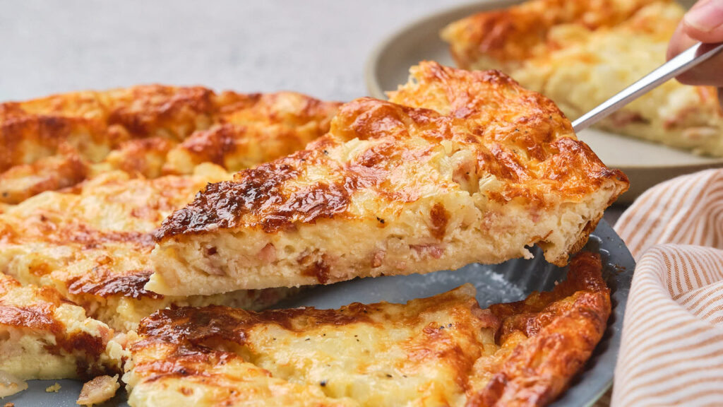 A hand lifts a slice of golden-brown, cheesy quiche from a whole pie resting on a blue plate.