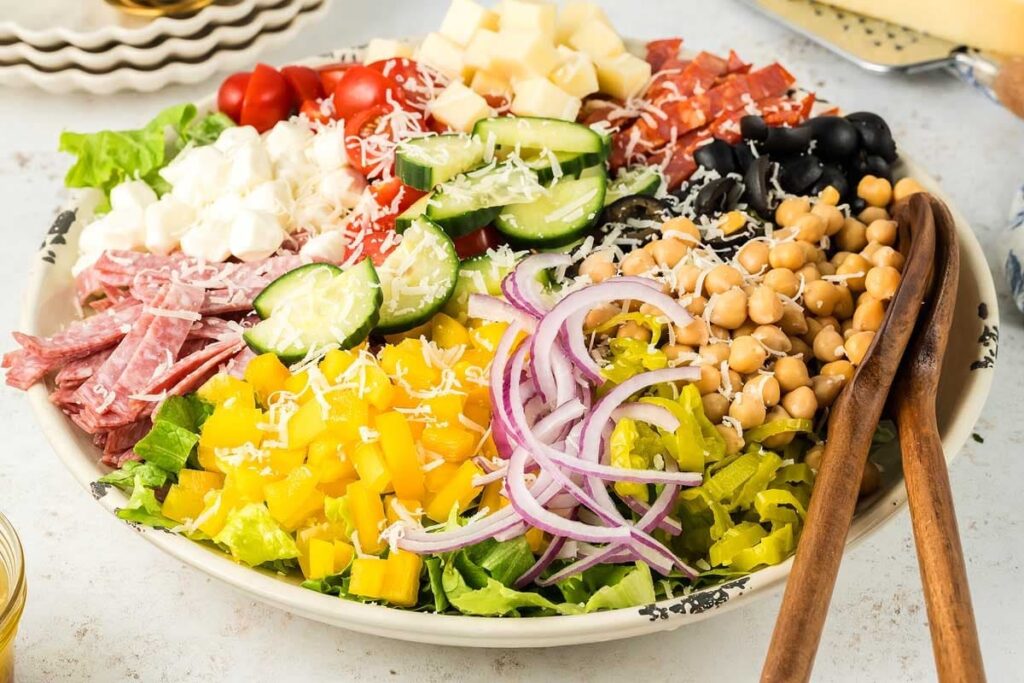 A colorful Italian salad with fresh veggies, cheese, chickpeas, pepperoni, and salami over a bed of lettuce.