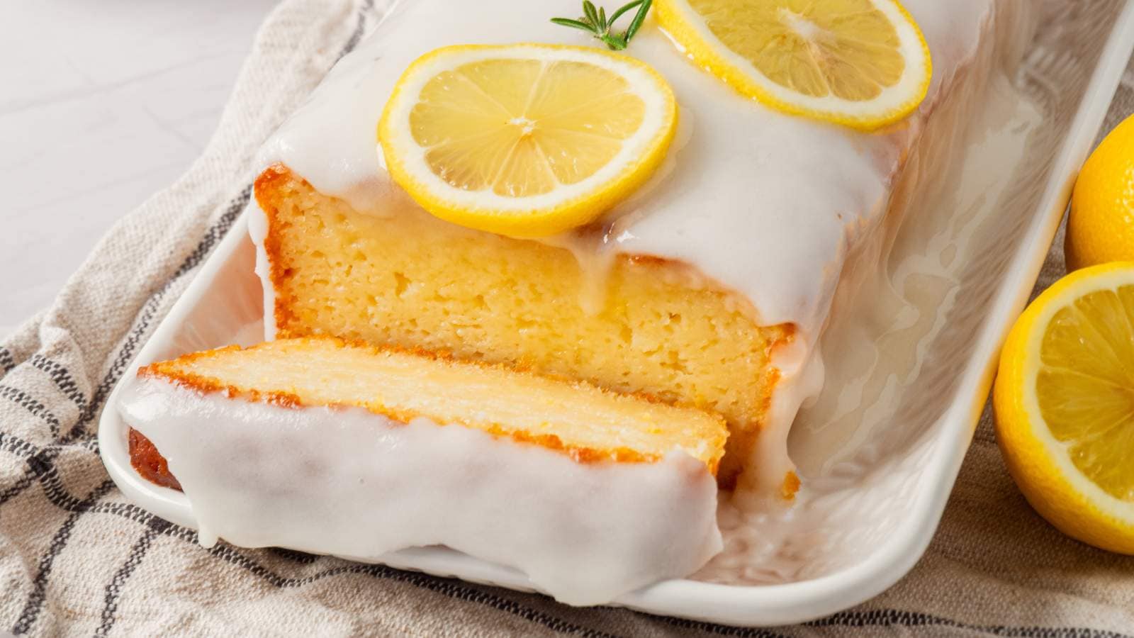 Lemon loaf cake on a white tray, topped with glaze and lemon slices, with a cut piece in the foreground and whole lemons nearby.