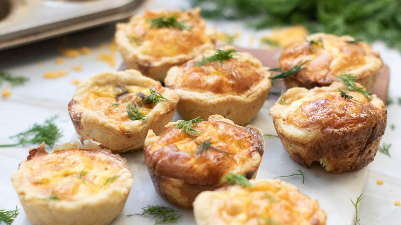 Mini salmon quiches arranged on a marble surface, topped with fresh dill, with a baking tray and more dill in the background.
