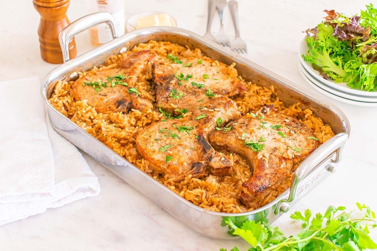 A baking dish with four cooked pork chops on top of seasoned rice, garnished with chopped herbs. Plates and a salad are in the background.