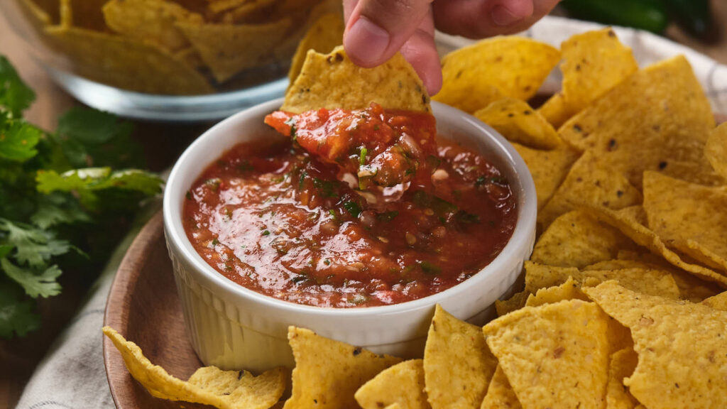 A person's hand dipping a piece of nacho chip in a salsa ranchera sauce.