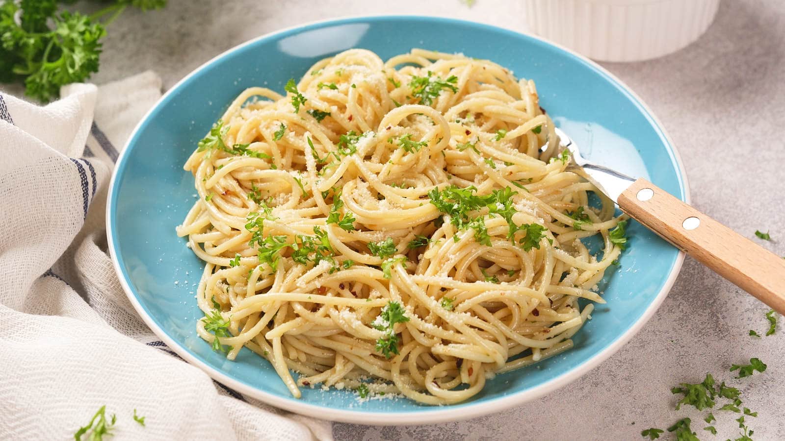 A blue plate of spaghetti garnished with chopped parsley and parmesan cheese, with a fork resting on the side.