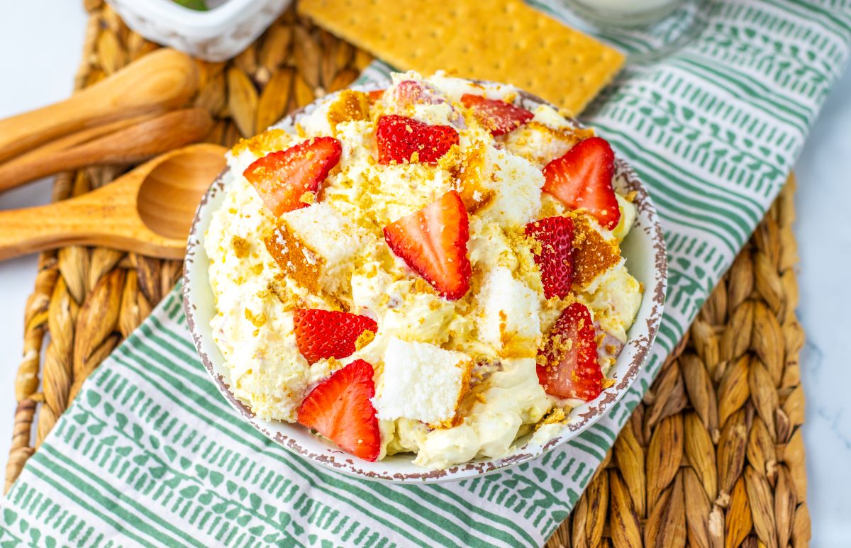 A bowl of dessert with whipped topping, strawberry slices, and crumbled graham crackers, placed on a patterned cloth with wooden spoons beside it.