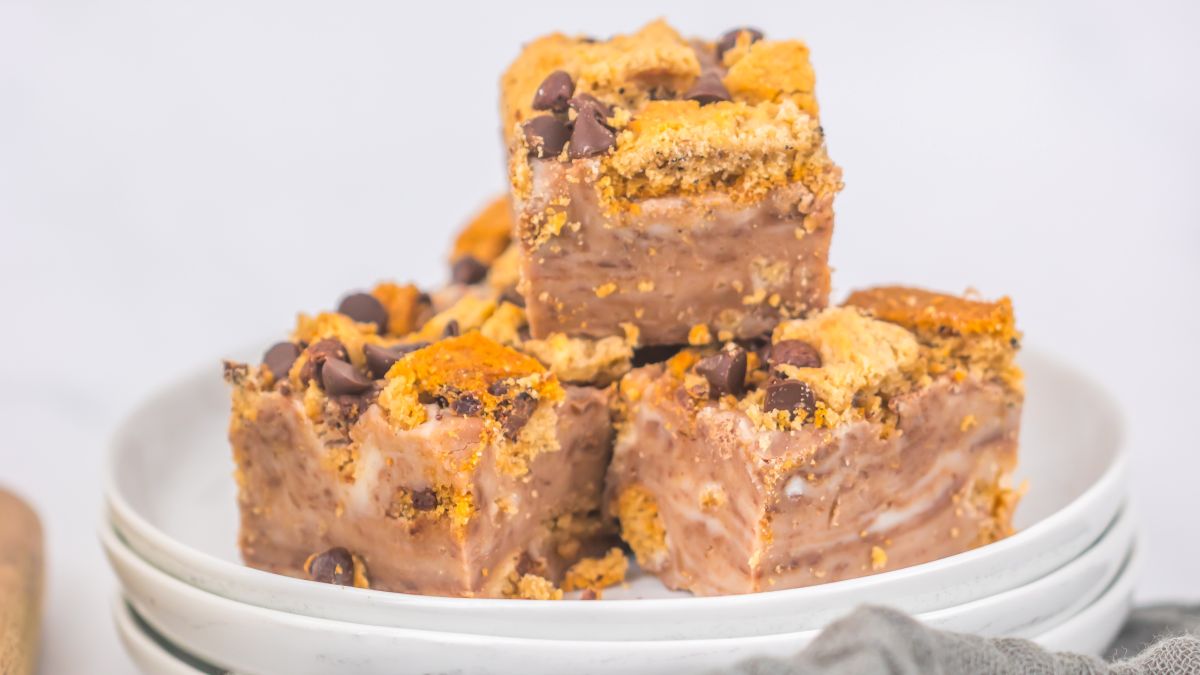 A plate stacked with fudge squares topped with crumbled cookies and chocolate chips, set on a gray cloth with a blurred background including a glass of milk and a bowl of white candies.