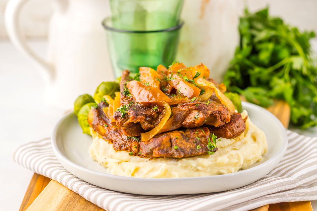 A plate of mashed potatoes topped with slices of cooked meat, sautéed onions, and herbs, served with Brussels sprouts.