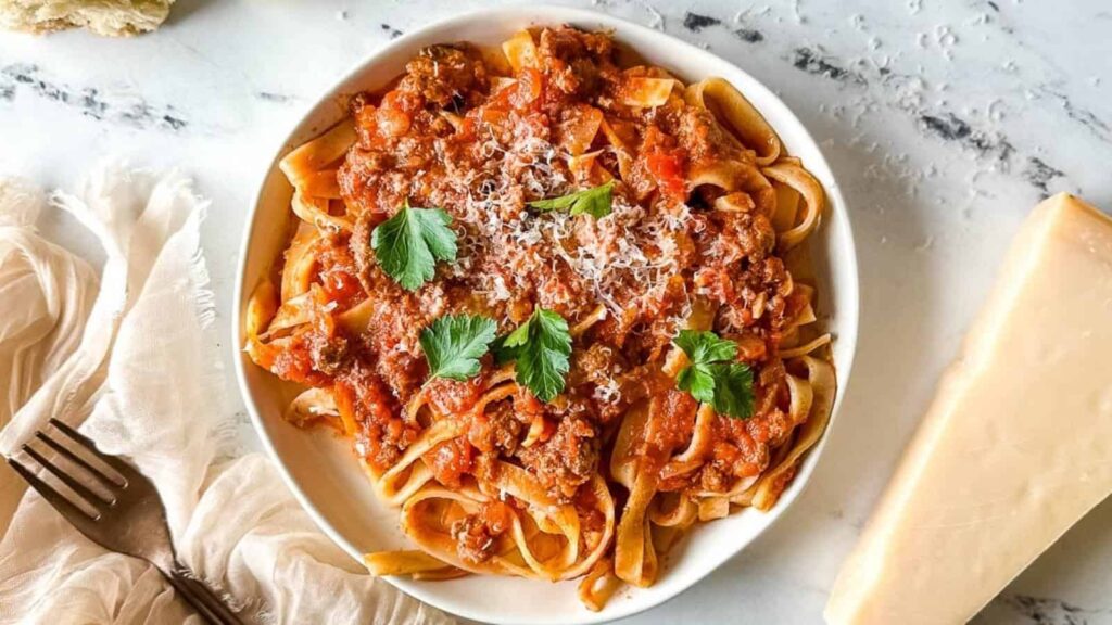 a plate of slow cooker lamb ragu over pappardelle pasta surrounded by a block of parmesan, a white linen, and torn baguette.